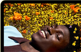 MacBook Neo, citrus color, screen shows a smiling person lying in a sunny field of flowers
